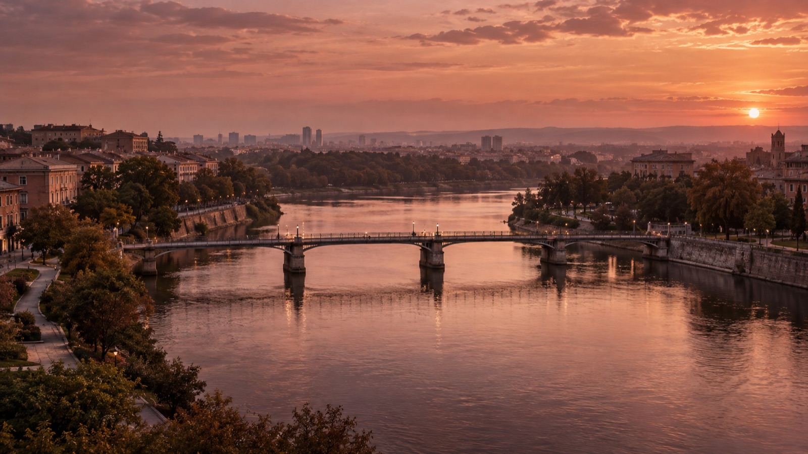 A bridge connecting two riverbanks, with public green space and a distant cityscape